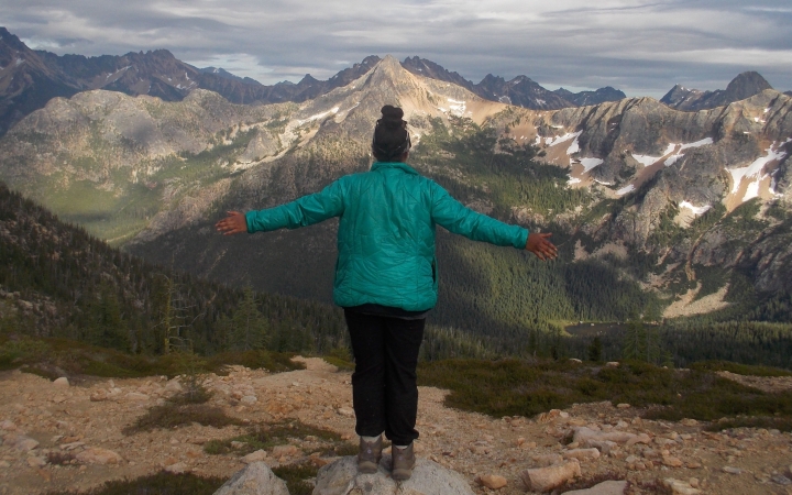 A person facing away from the camera spreads their arms out wide while standing on a rock overlooking a vast mountainous landscape.